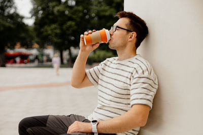 Young adult man sitting outdoors, drinking coffee, enjoying good weather and city view. 