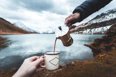 Cropped hand holding coffee cup while person pouring drink against sea