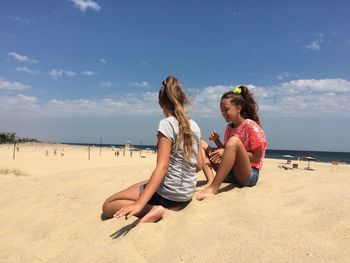 Teenage female friends relaxing at sandy beach against sky on sunny day