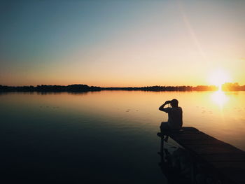 Man sitting on lake against sky during sunset