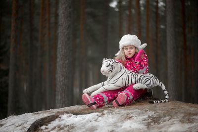 Girl in warm clothing gesturing while sitting with toy tiger by trees in forest