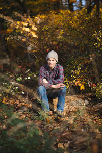 Confident teen boy in plaid shirt and jeans sits on log in woods.