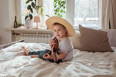 Little toddler boy in hat playing ukulele guitar at home, rustic style. lifestyle concept