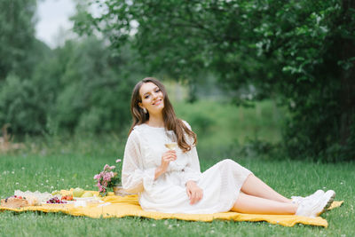 A happy smiling girl holds a glass of juice or wine and sits on a yellow blanket on a summer picnic 