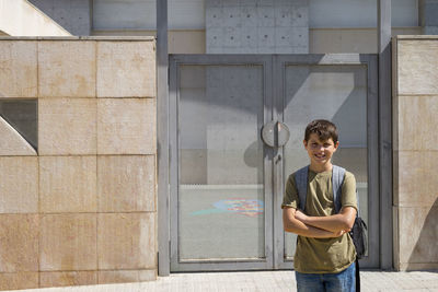 Portrait of smiling boy with backpack standing against metal door during sunny day