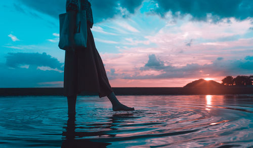 Low section of woman walking in sea against sky during sunset