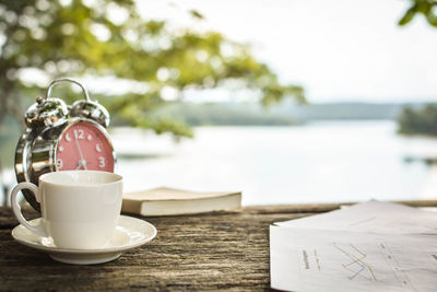 Coffee cup with alarm clock and book on table against lake