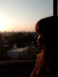 Close-up of woman looking at city through window during sunset