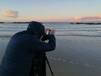 Photographer wearing hooded jacket photographing through tripod camera at beach