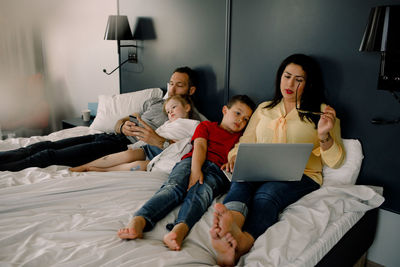 Parents using technologies while sitting with children on bed in bedroom