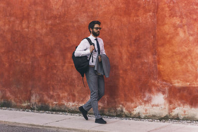 Full length of businessman with backpack walking on sidewalk against wall in city