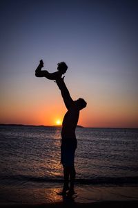 Silhouette father tossing child at beach against clear sky during sunset