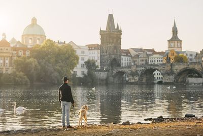 Rear view of man with dog by river against buildings