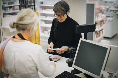 High angle view of senior customer holding prescription paper with female owner standing at checkout in pharmacy store