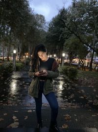Young woman using phone while standing at park during dusk