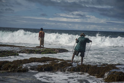 Rear view of men standing waves at beach against sky