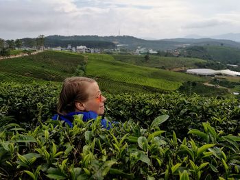 Young woman wearing sunglasses lying amidst tea crops at farm against cloudy sky