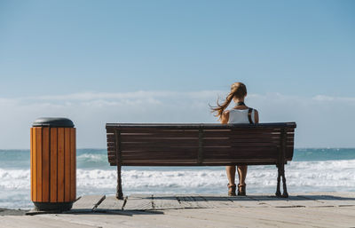 Young beautiful woman, with hair blowing in the wind, sitting on bench, looking to the blue ocean. 