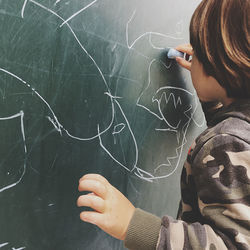 Rear view of boy drawing on blackboard at classroom