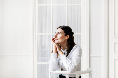 Young woman looking away while standing against window at home