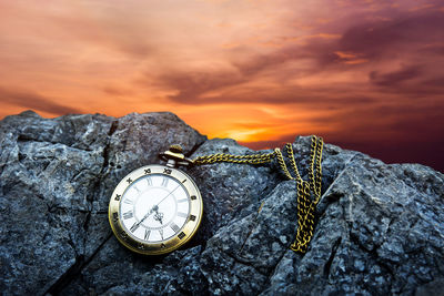 Close-up of pocket watch on rock against sky during sunset