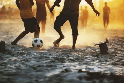 Low section of friends playing soccer at beach during sunset