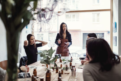 Smiling female owner standing with perfume communicating with multi-ethnic colleagues at workshop