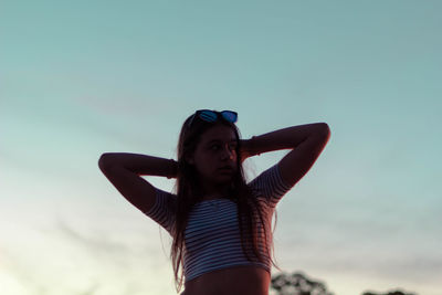 Low angle view of young woman with hands behind head standing against sky during sunset