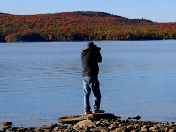 Rear view of man standing by lake taking pictures. 