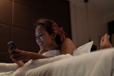 Smiling young woman using mobile phone while lying on bed