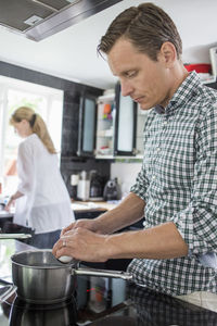 Husband cooking while wife cleaning utensils in background at kitchen
