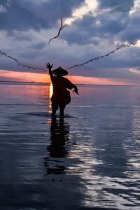 Silhouette fisherman throwing fishing net on sea against sky during sunset