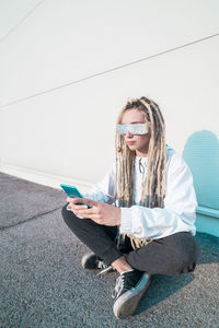 Young woman using mobile phone while sitting on road against wall