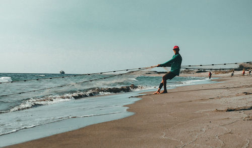 Full length of man fishing while standing on beach against clear sky