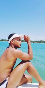 Shirtless man sitting at beach against blue sky