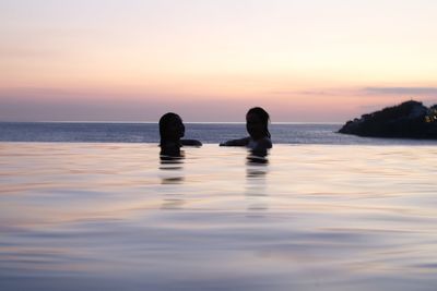 Silhouette people swimming in sea against sky during sunset