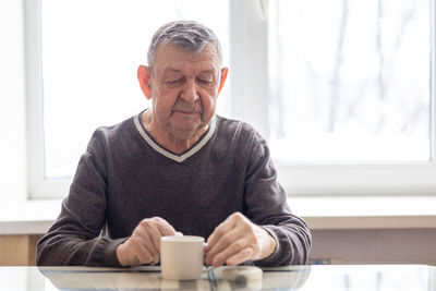 Sad portrait of elderly man. the senior sits at table, thoughtfully looking at mug of water 