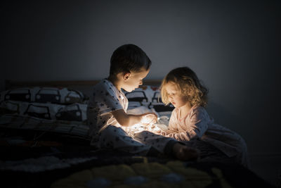 Siblings holding illuminated string light while sitting on bed against wall at home