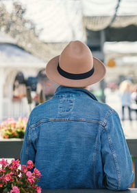 A man in a denim jacket and a hat stands with his back and looks at the busy street. 