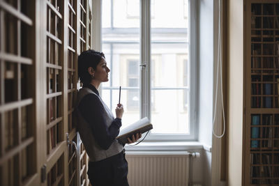 Profile view of thoughtful lawyer holding book while leaning on shelf in library