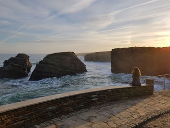 Rear view of woman sitting on wall over sea against sky during sunset