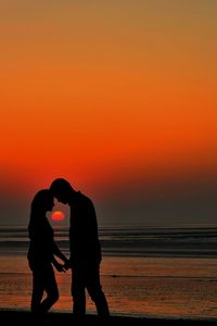 Silhouette couple cuddling at beach against orange sky