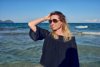 Smiling mature woman with hand in hair standing at beach against clear sky