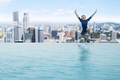 Happy boy with arms raised jumping in infinity pool against cityscape