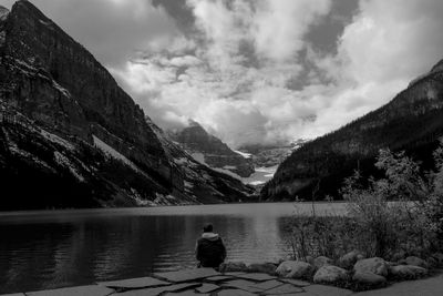Rear view of man sitting by lake against cloudy sky