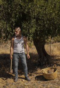 Adult man in white tank top and jeans standing with olive tree in summer