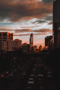 Traffic on city street by buildings against sky during sunset