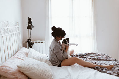 Side view of young woman playing with dog while sitting on bed at home