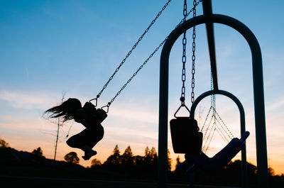 Silhouette girl swinging at playground against sky during dusk