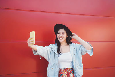 Smiling young woman taking selfie while standing by red wall